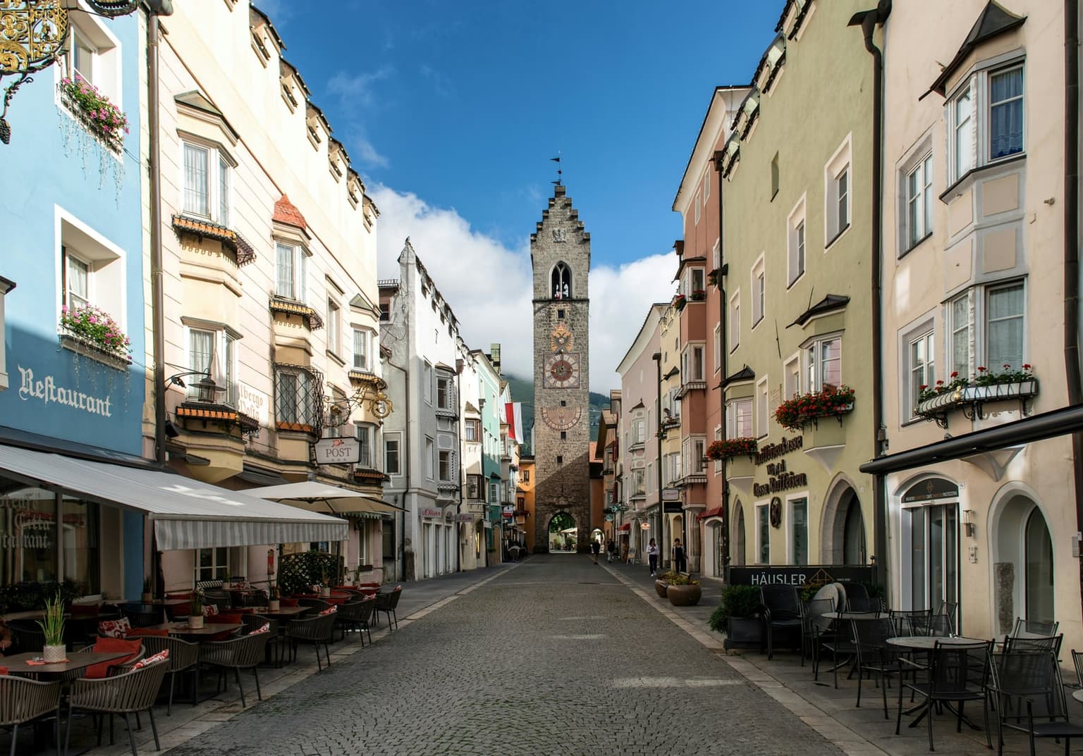 Vipiteno's stepped-gable Zwölferturm clock tower at the end of the cobbled main street, lined with pastel houses with bay windows, flower boxes and cafe tables