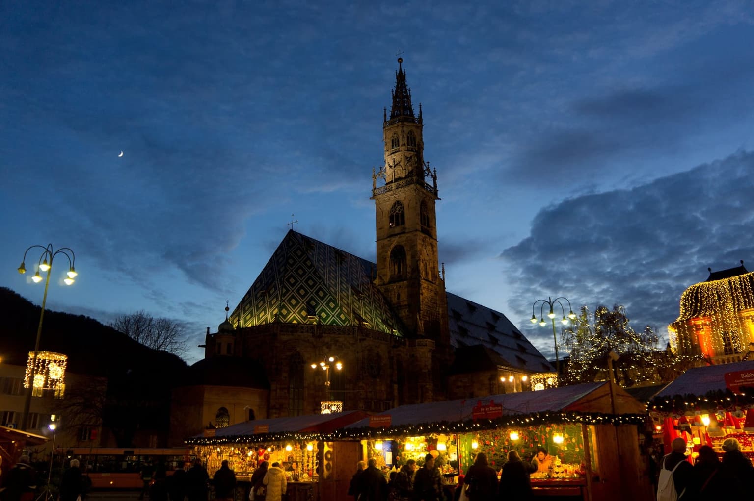 Bolzano cathedral, with its green and gold diamond-patterned roof and tall spire, at dusk above a row of lit wooden Christmas market stalls draped in garlands, with shoppers in front
