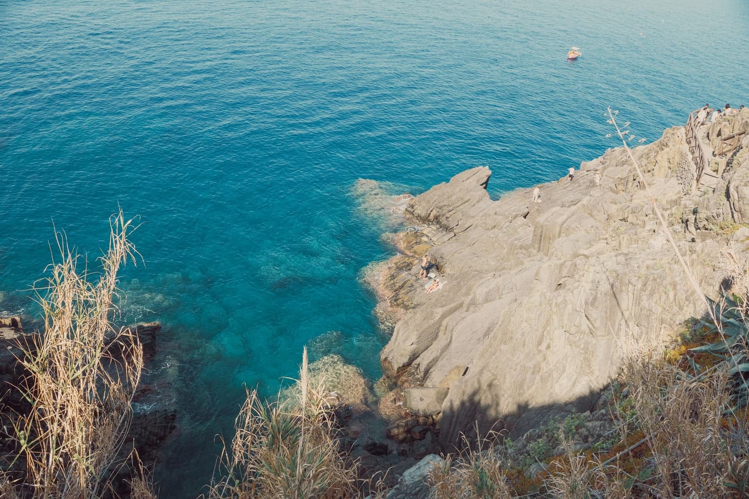 View from a cliff down onto a rocky point on the Cinque Terre coast, with clear turquoise water below, a few people on the rock ledge, and a small boat far out on calm sea