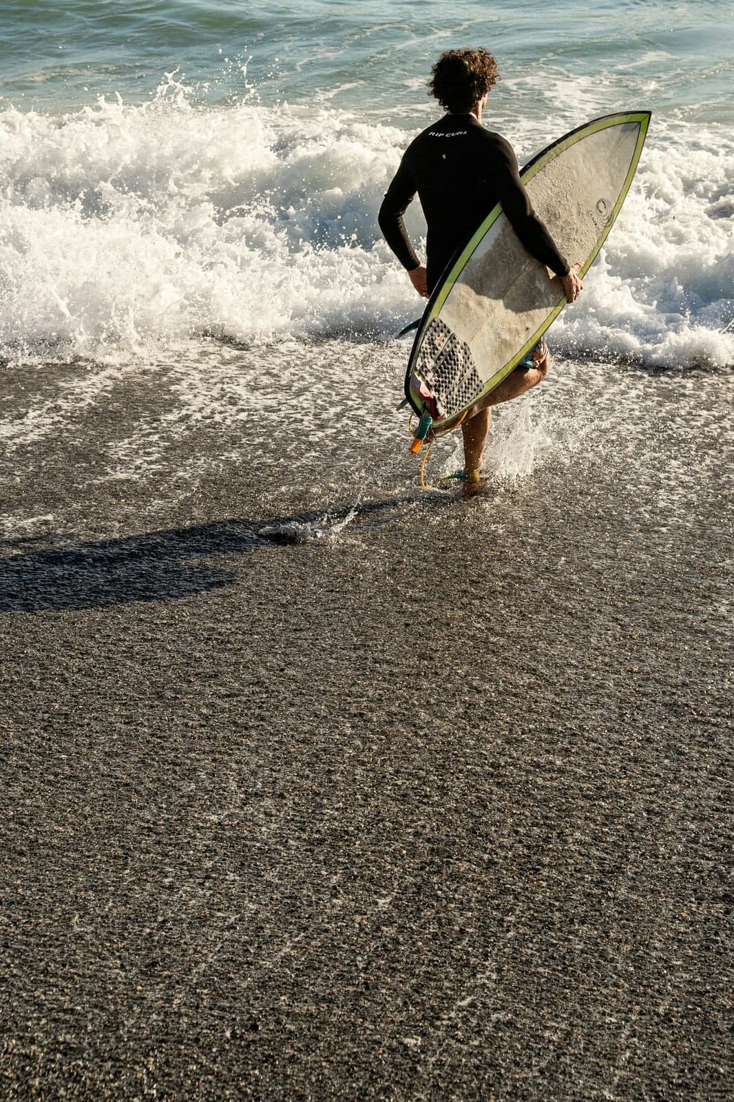 A surfer in a black wetsuit carrying a board across dark gray sand toward breaking waves