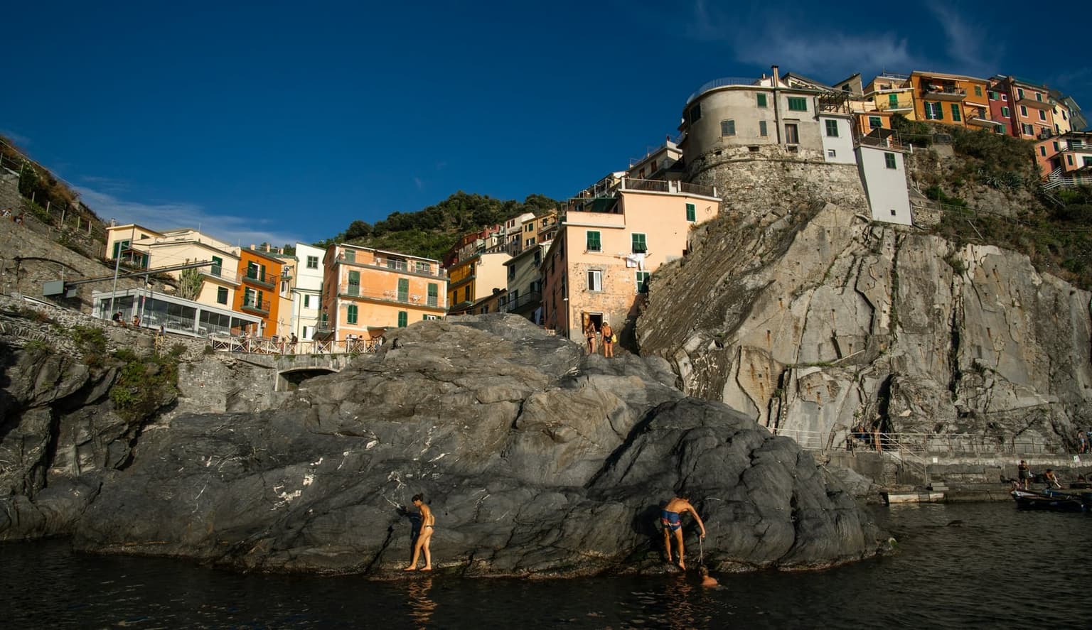 Two swimmers climbing onto a large gray rock slab at the water's edge, with pastel-colored houses stacked on the cliff above