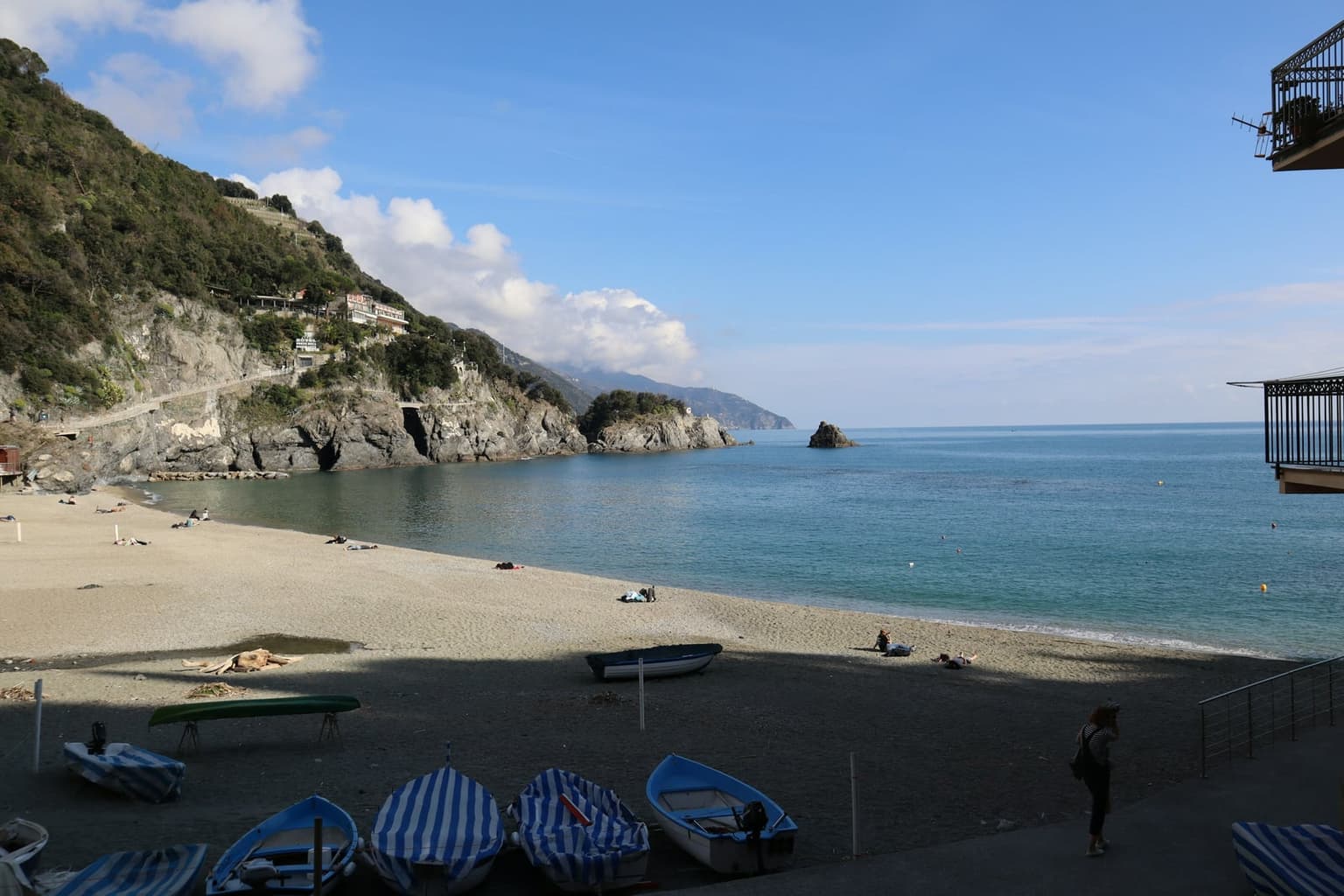 Monterosso old-town beach with small boats pulled up on the dark sand in the foreground, a few sunbathers, calm turquoise water, and the coastal path running along the rocky headland