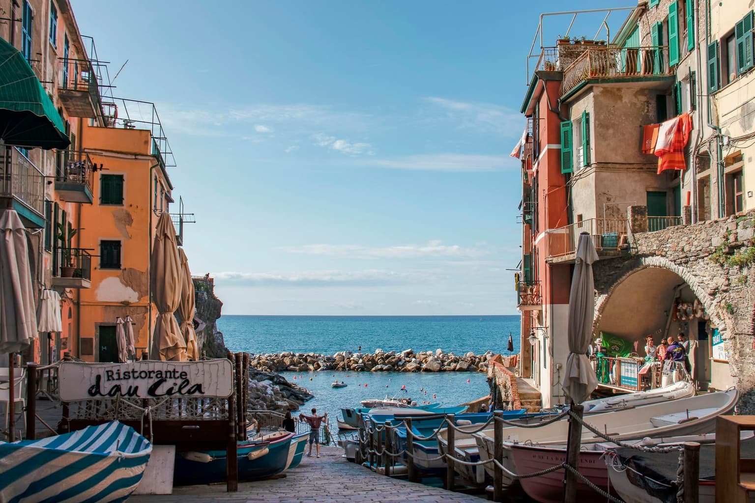The slipway at Riomaggiore marina with small boats pulled up behind rope barriers, a breakwater of rocks and calm blue sea beyond, framed by orange and red houses