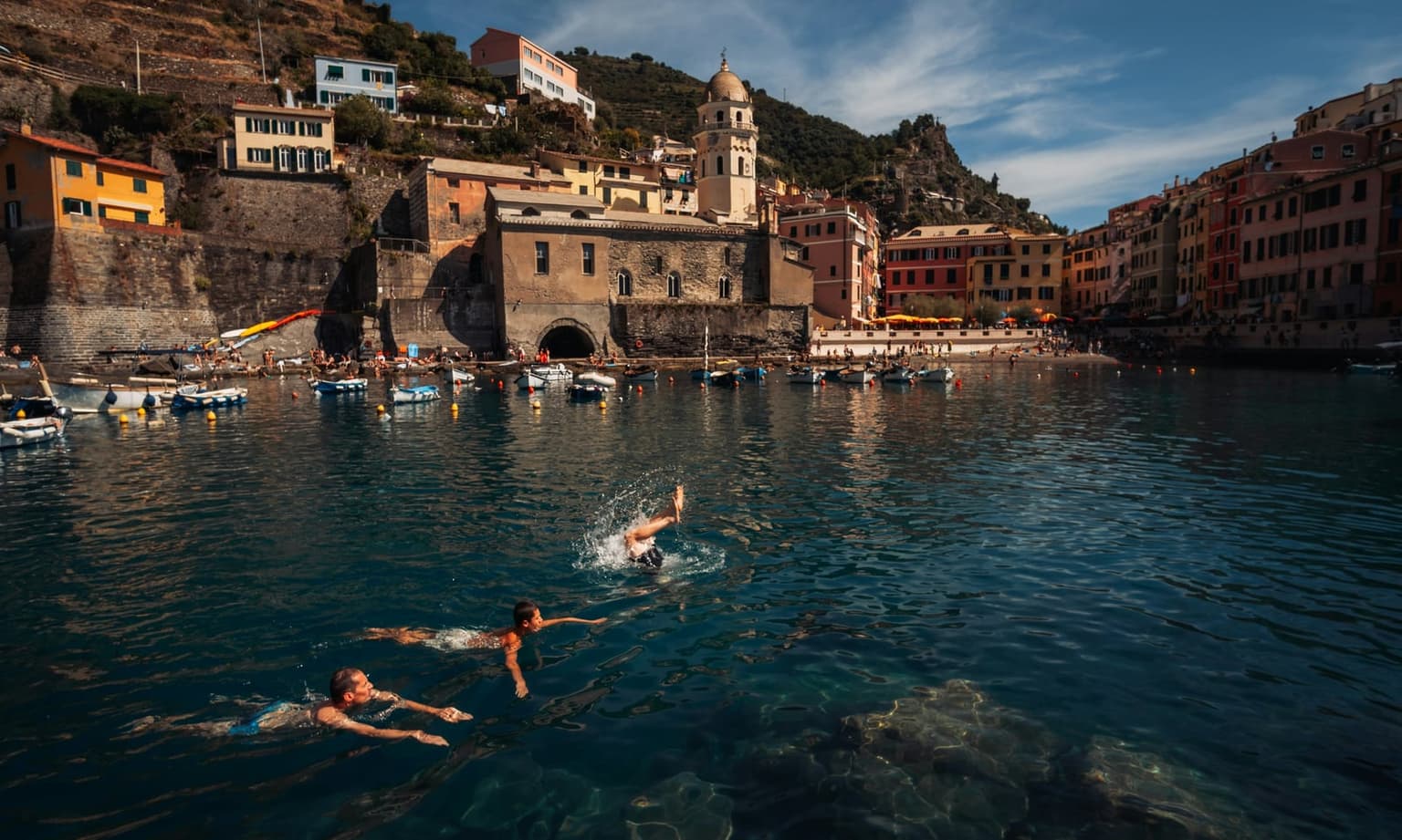 Three people swimming in the dark blue harbor of Vernazza, one mid-dive with a splash, with moored small boats, the stone harbor wall, the church bell tower and rows of colored houses behind