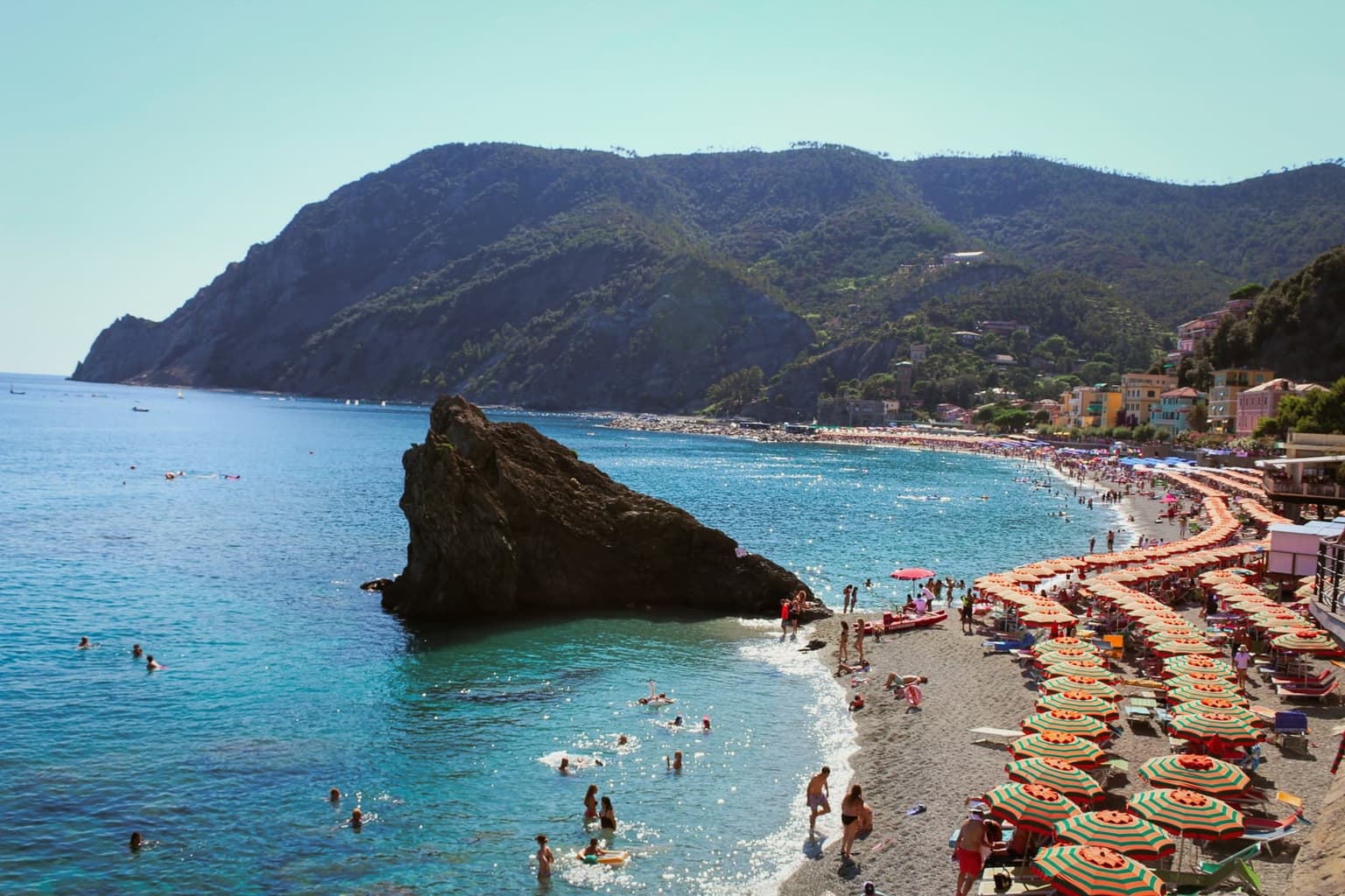 Fegina beach at Monterosso from above, with rows of orange and green striped umbrellas on the pebbly sand, swimmers in the clear blue shallows, and a large dark rock offshore