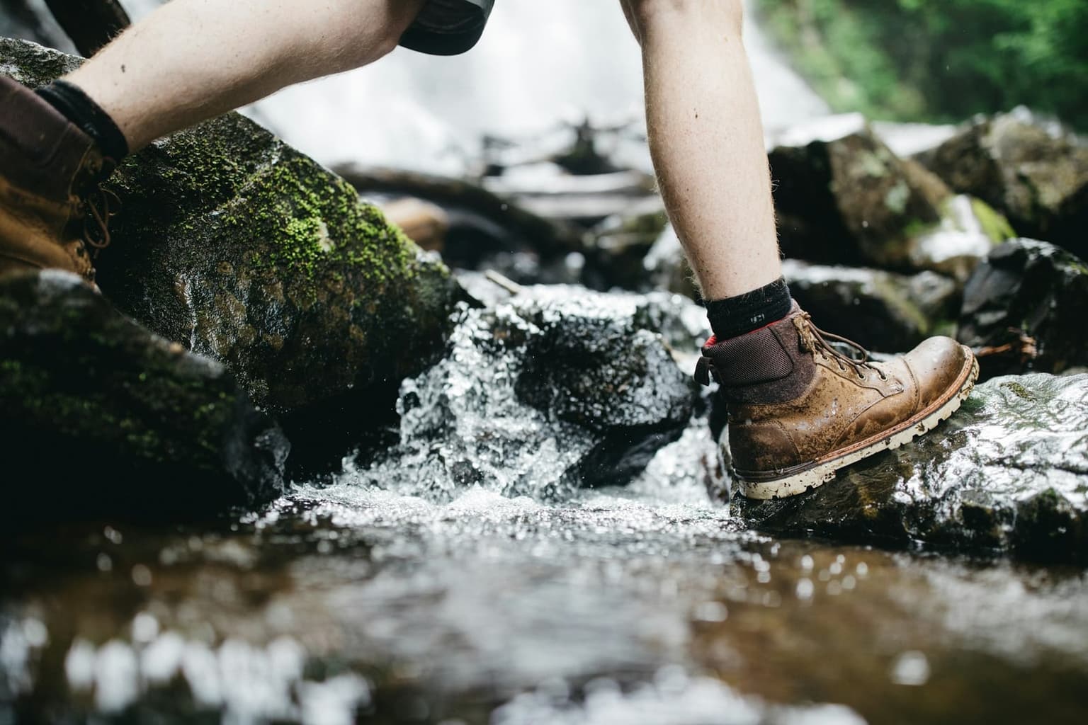 A hiker's leather boot with a lugged sole steps onto wet rock beside running water.