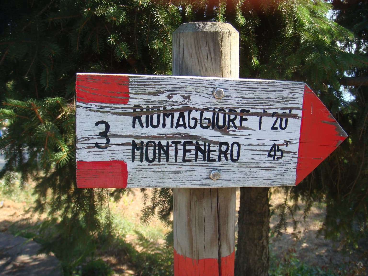 A wooden trail sign with red and white ends, the number 3, and arrows pointing to Riomaggiore and Montenero.