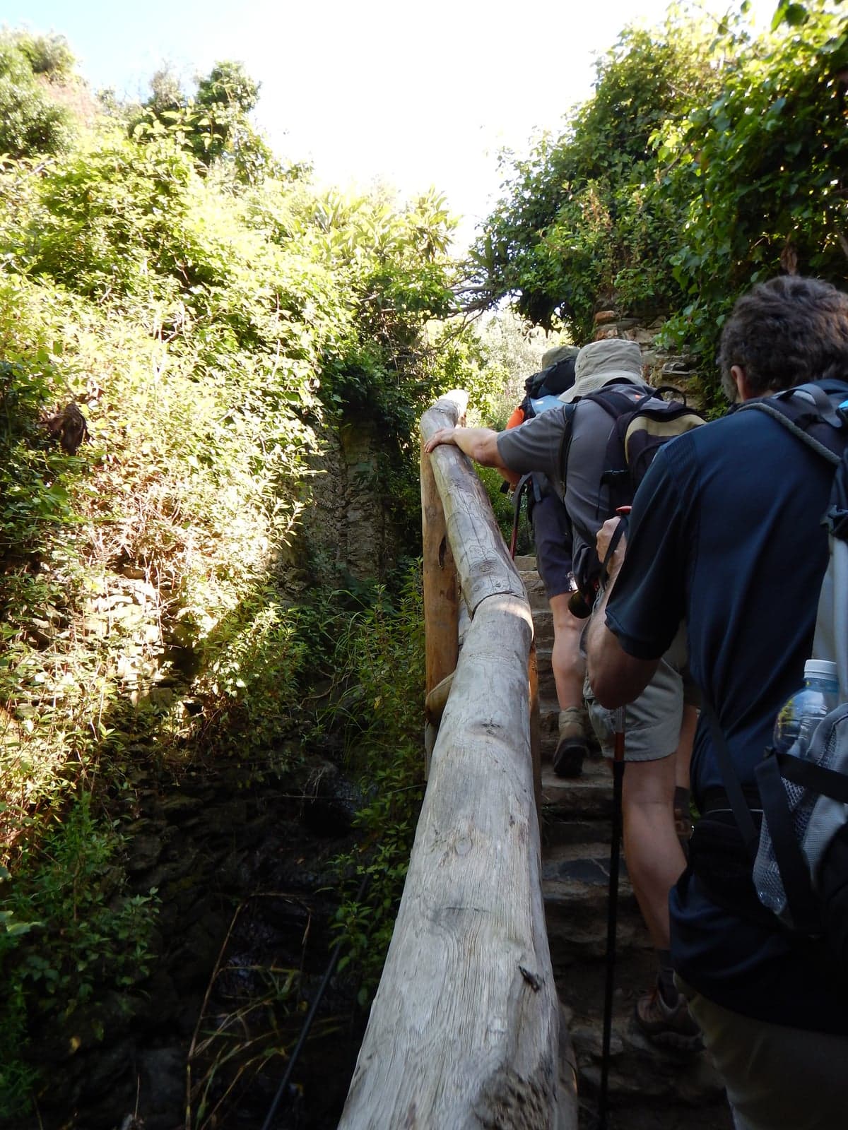 Hikers with backpacks and trekking poles climb a steep stone stair beside a wooden handrail on the coastal trail near Monterosso.