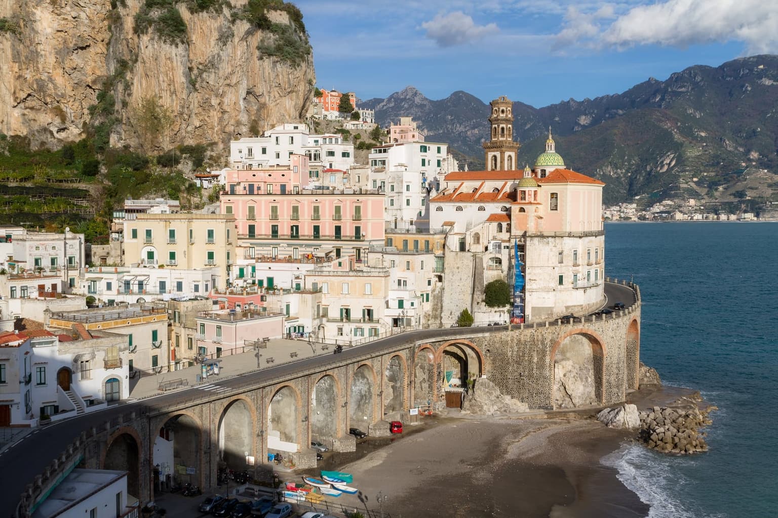 Atrani's white and pink houses stacked beneath a sheer cliff above its small dark-sand beach, the coast road crossing the village on stone arches, and a church with a tiled dome and bell tower on the rock above the sea