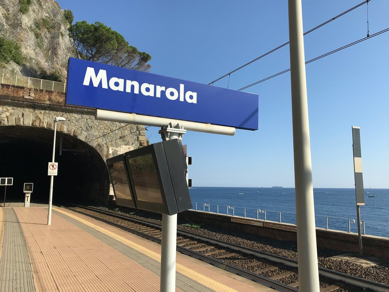 The blue Manarola station sign on the platform, with a tunnel mouth beside it and the sea beyond the tracks