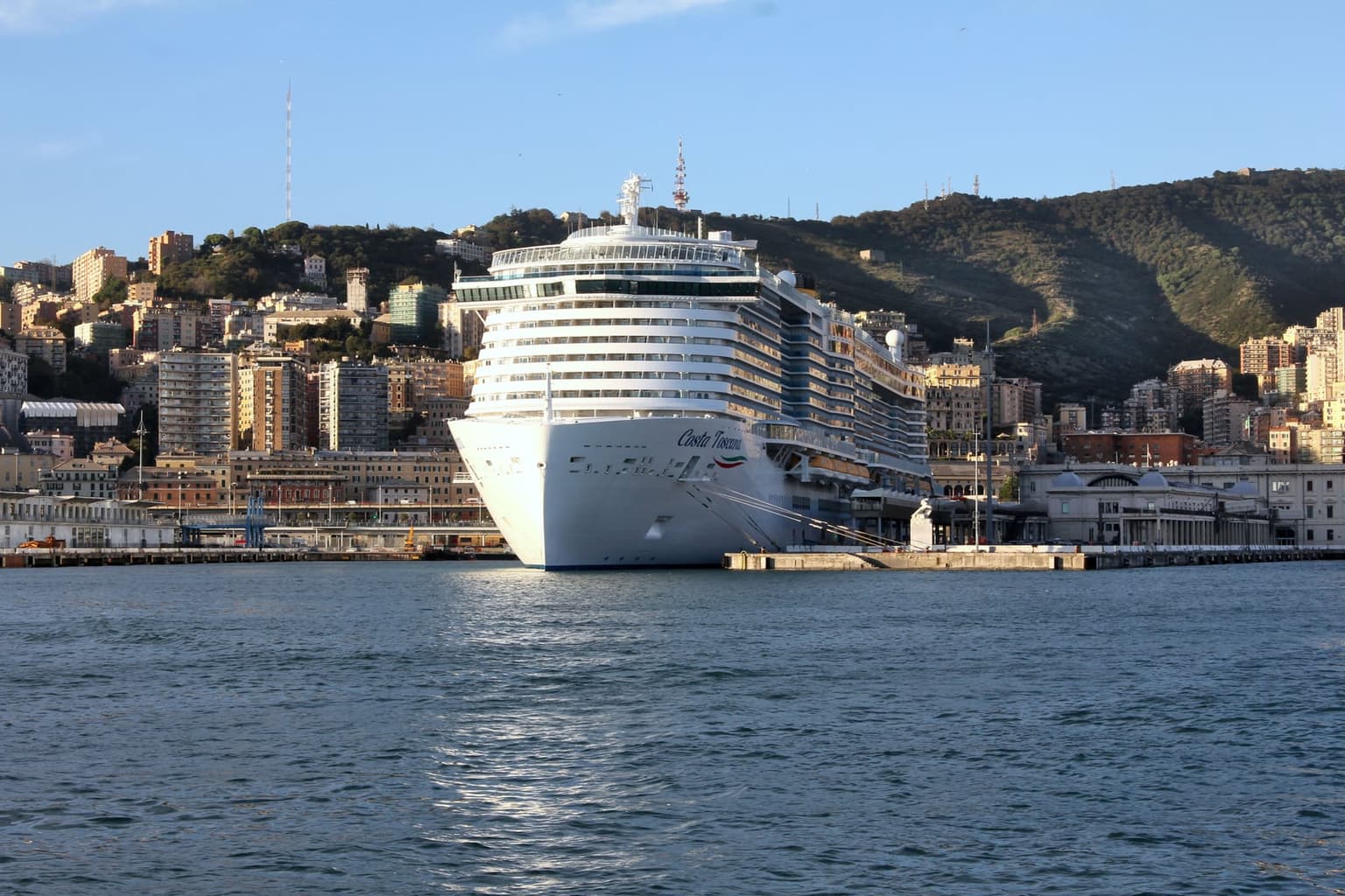 The Costa Toscana cruise ship docked bow-on at Genoa's port, with the city's hillside apartment blocks and a wooded hill rising directly behind it under a clear sky.