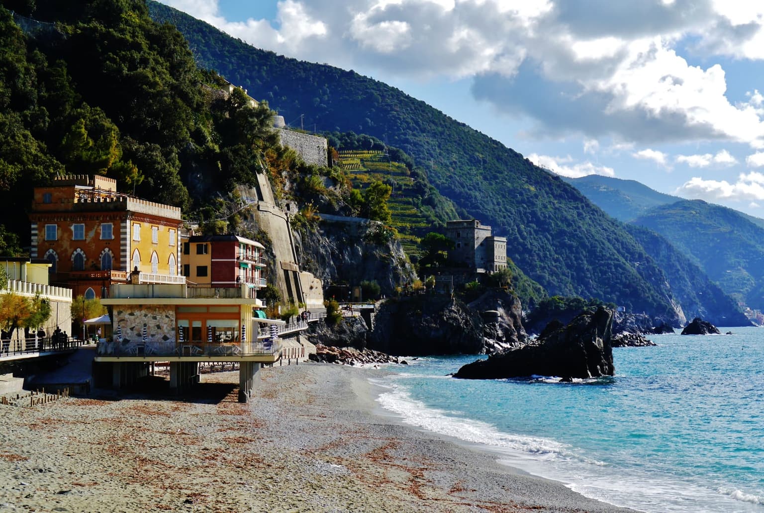 Monterosso al Mare's beach at ground level, looking toward the old town's waterfront buildings, with a fortified tower on the rocky point behind.