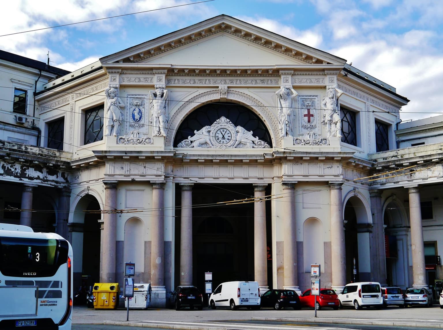 Close view of the Genova Piazza Principe station facade: a neoclassical stone front with a central clock and flanking telamon (Atlas) statues, under a clear blue sky.