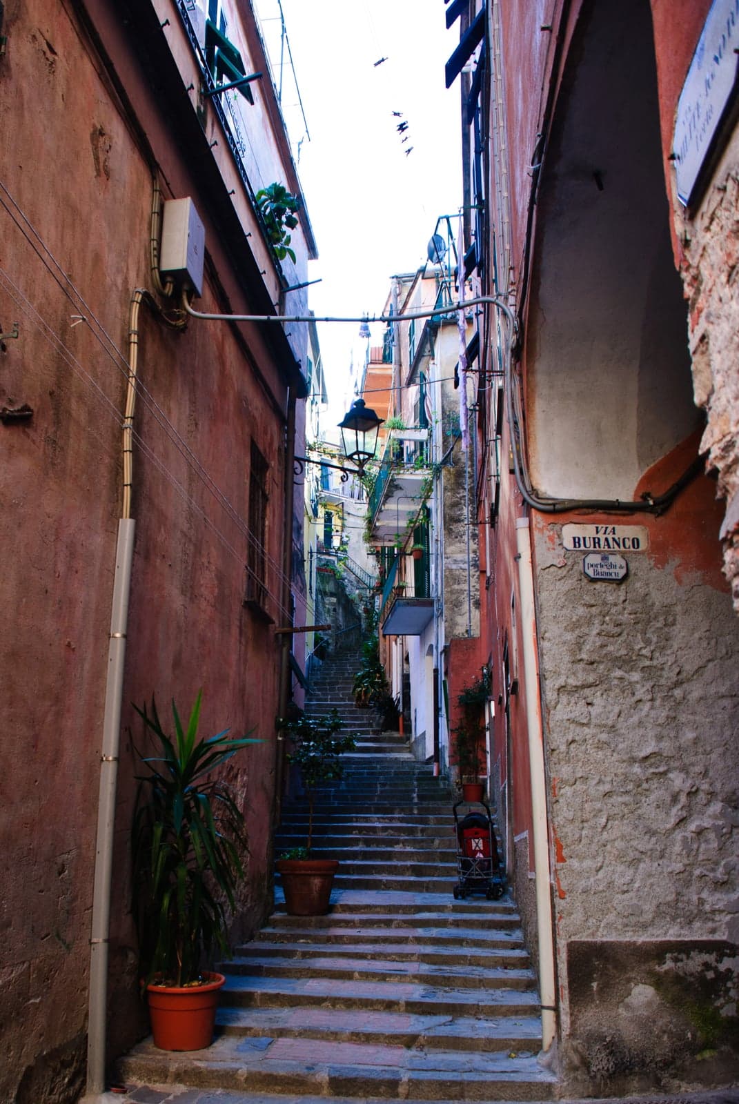 Via Buranco in Monterosso al Mare, a narrow stepped lane climbing steeply between weathered red houses, with potted plants on the steps and a street lamp overhead