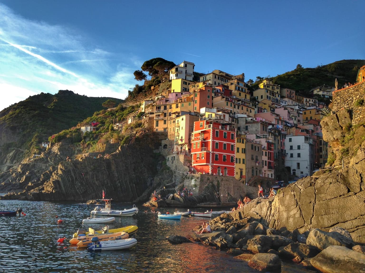 Riomaggiore's harbor in warm late-afternoon light, small boats moored below the steep cluster of colorful houses and people sitting on the rocks at the water's edge