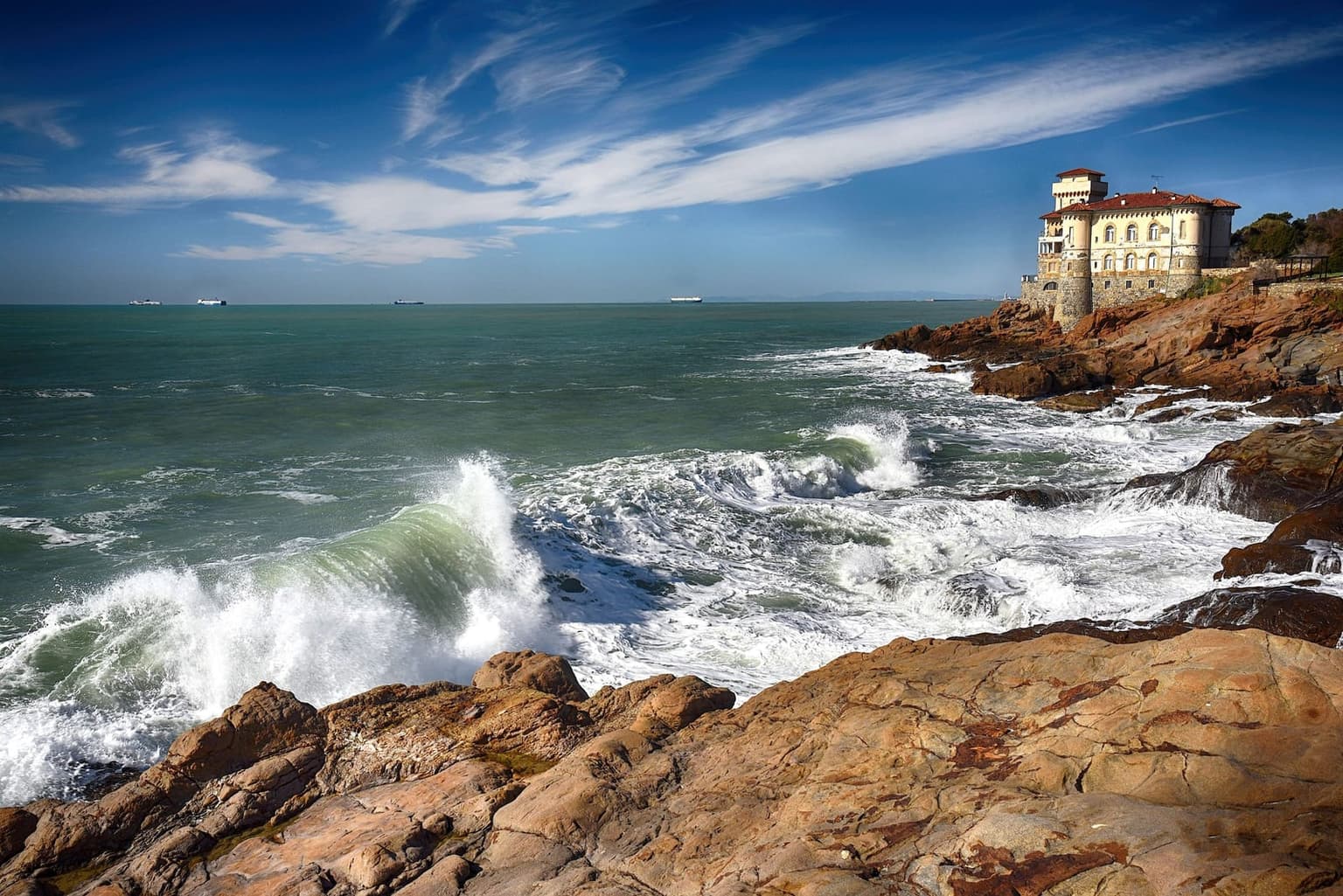 Waves breaking on reddish rocks on the Tuscan coast south of Livorno, with the Castello del Boccale villa on the headland