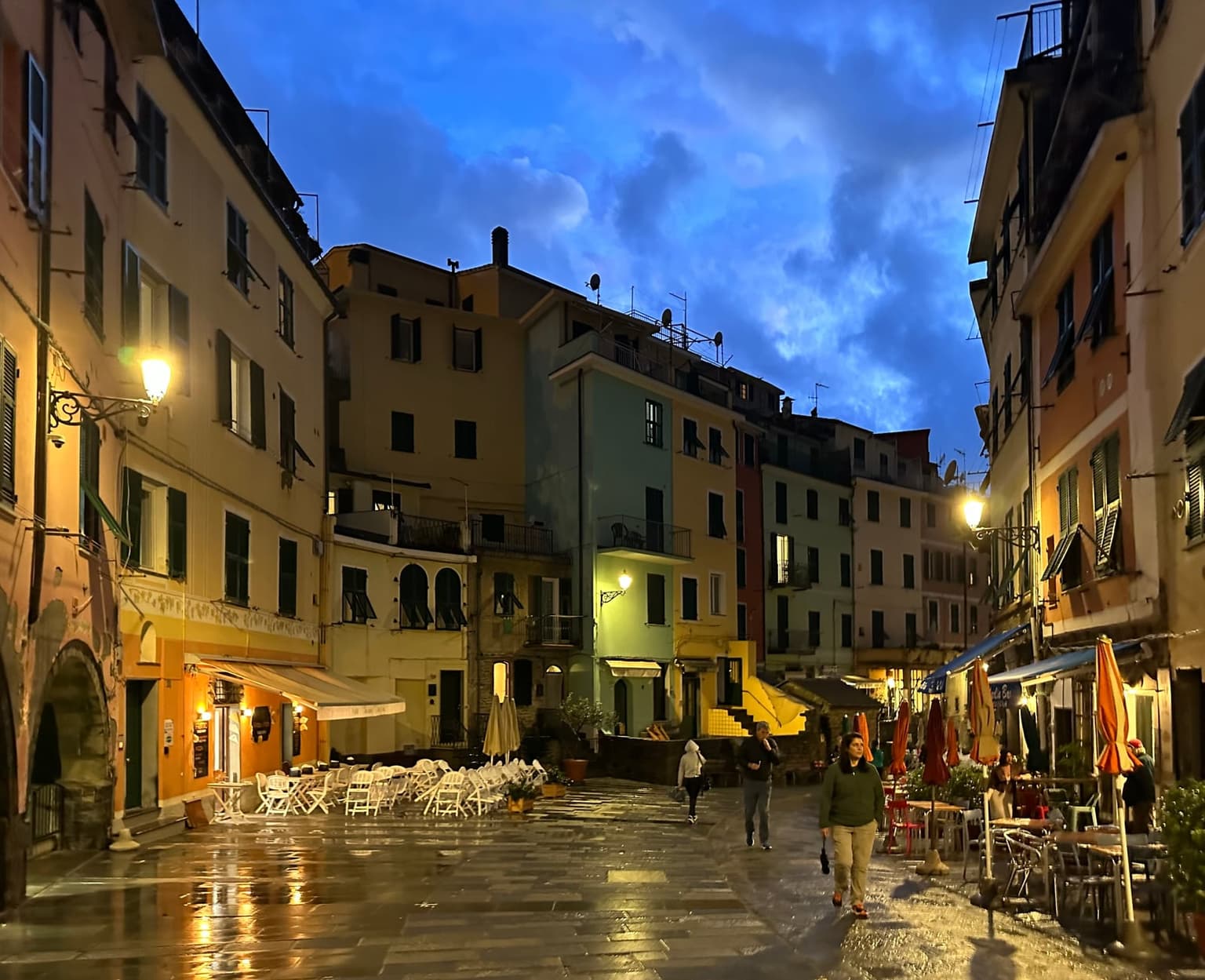Vernazza's main square at blue hour after rain, street lamps reflected in the wet paving, empty cafe chairs and only a few people walking
