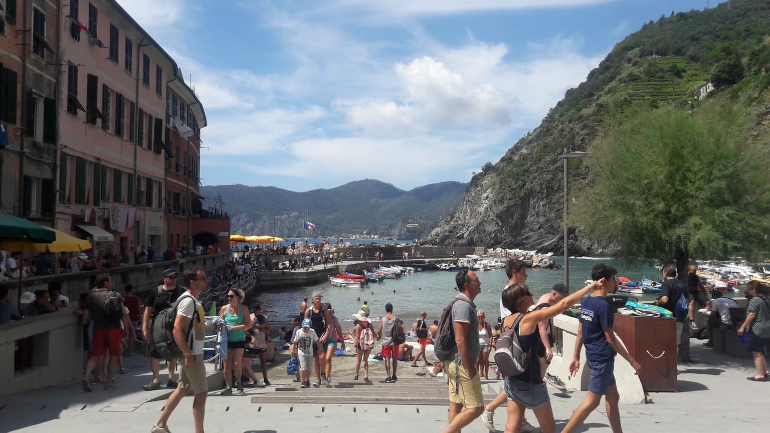Crowds of day-trippers with backpacks in Vernazza's harbor square on a sunny day, with the small beach, moored boats and breakwater behind them