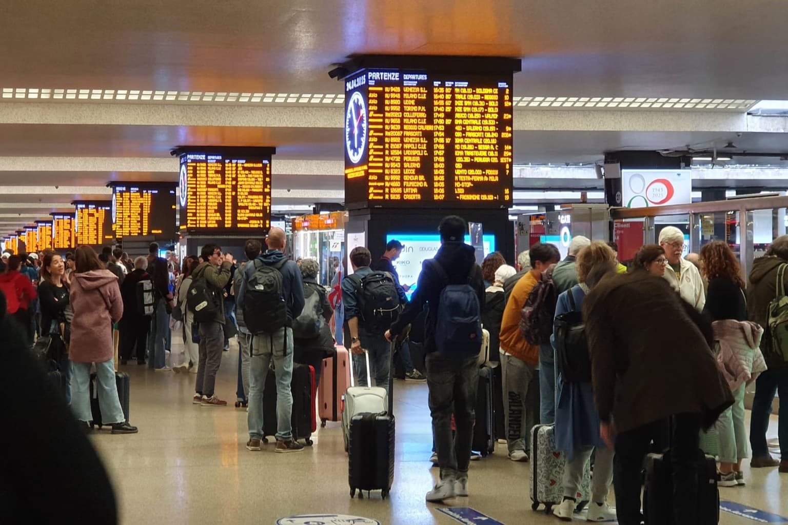 Travelers with suitcases and backpacks waiting under the departure boards in the main concourse of Roma Termini