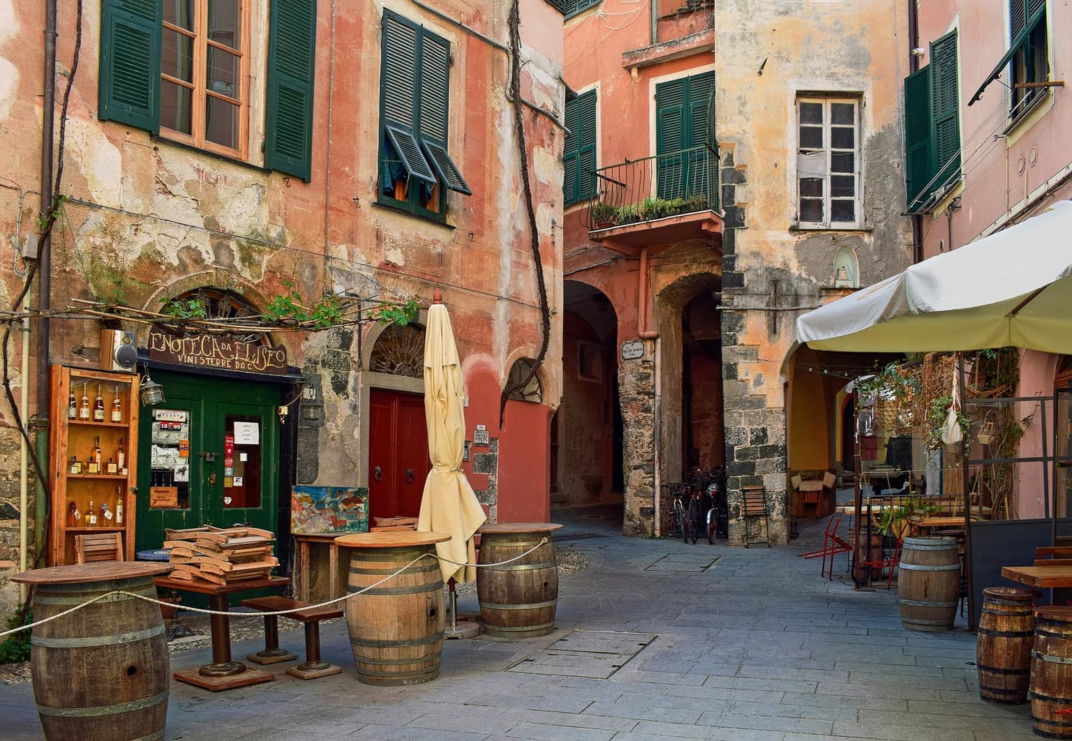 A small square in Monterosso al Mare's old town, with the Enoteca da Eliseo wine shop, wine-barrel tables and faded pink facades with green shutters, an arched passage leading off behind