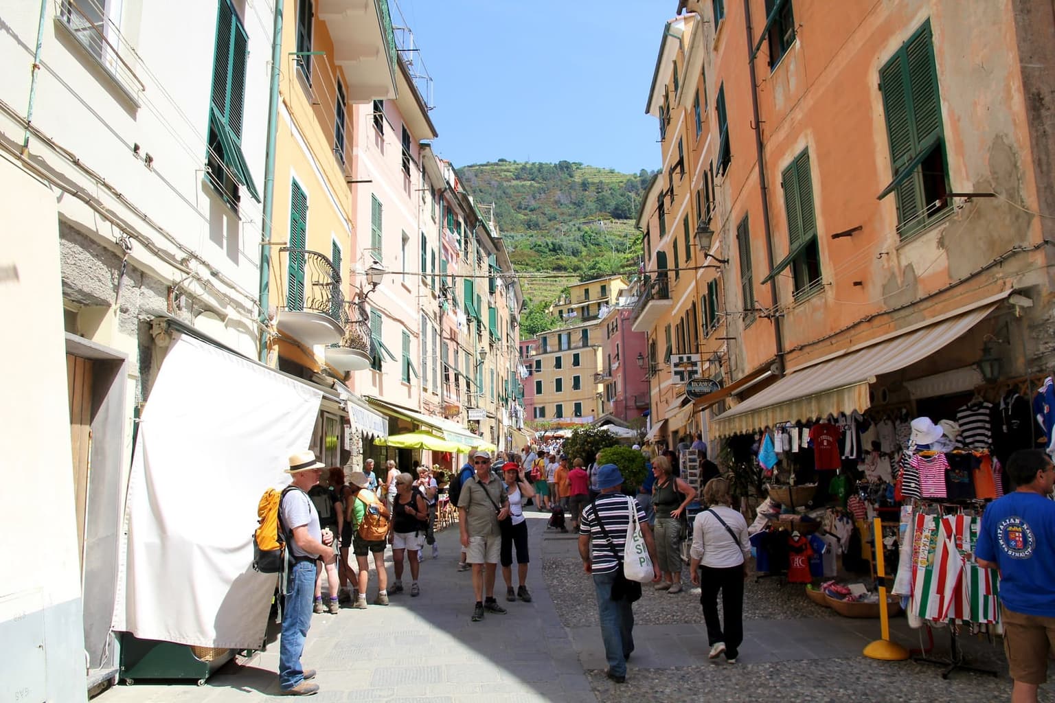 Vernazza's car-free main street on a sunny day, pastel houses with green shutters on both sides, souvenir stalls and hikers on foot, and a terraced green hillside rising beyond the rooftops