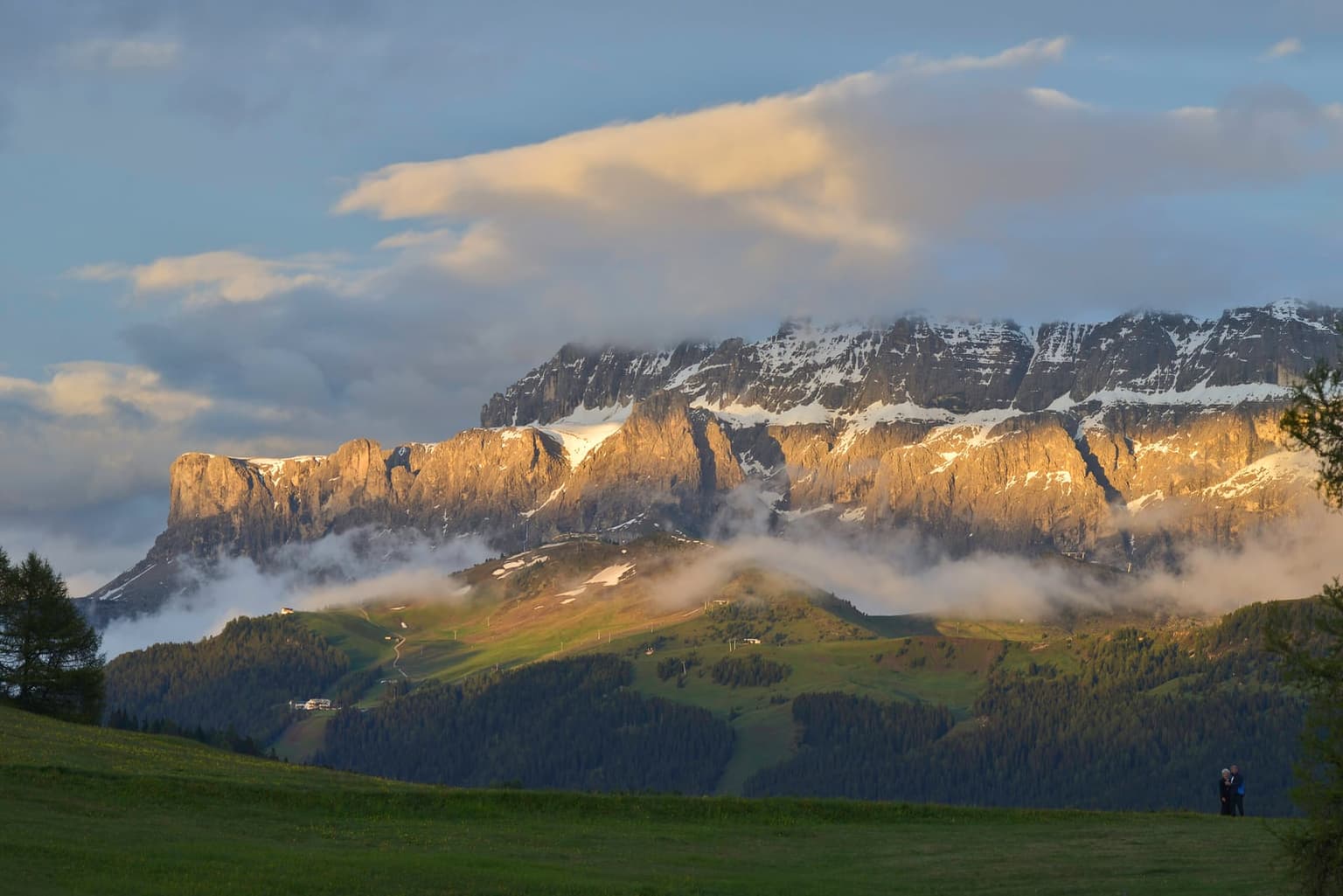A band of cloud clinging to the mountainside above Alpe di Siusi at evening, the snow-streaked peak lit gold above the cloud line, a clear green meadow in the foreground