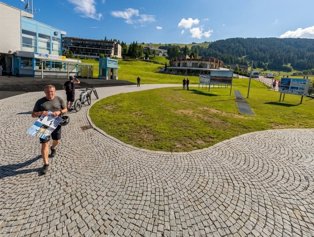 The Compatsch hub at the top of the Alpe di Siusi cable car: a lift station and ticket building beside a paved plaza, hotel buildings and forested hills in the distance, open meadow beginning just past the pavement