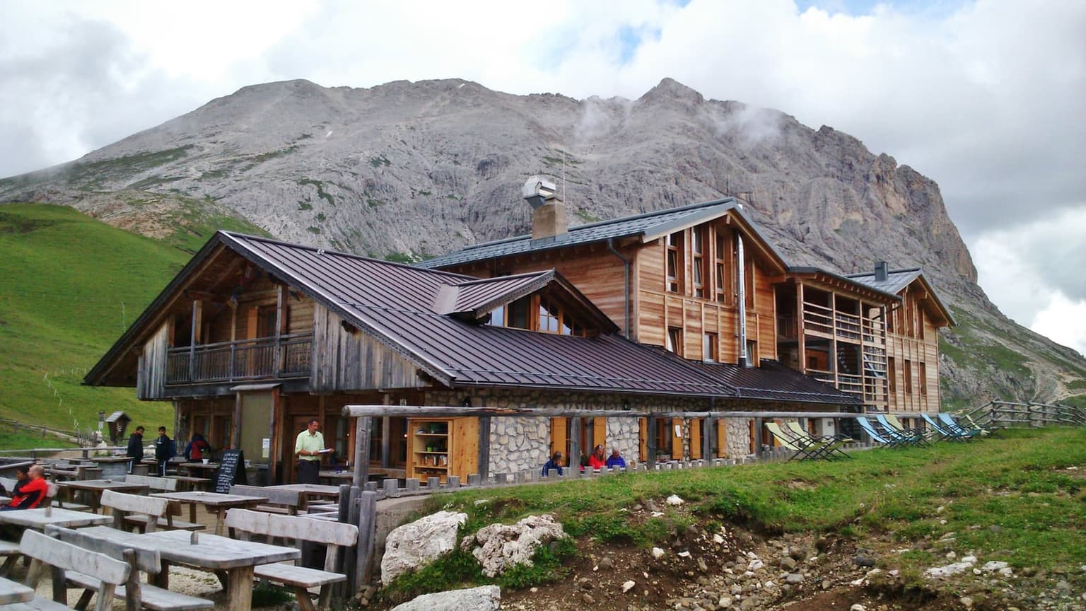 The Plattkofelhütte's sun terrace below Sassopiatto, wooden picnic tables and deck chairs on the grass, hikers and a waiter on the porch, the bare gray massif rising directly behind the hut