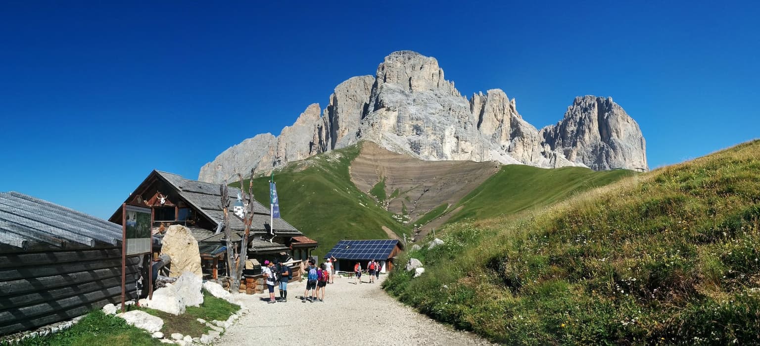Hikers on the gravel path to Rifugio Friedrich August, the sheer gray wall of Sassolungo rising directly behind the hut with a scree slope at its base