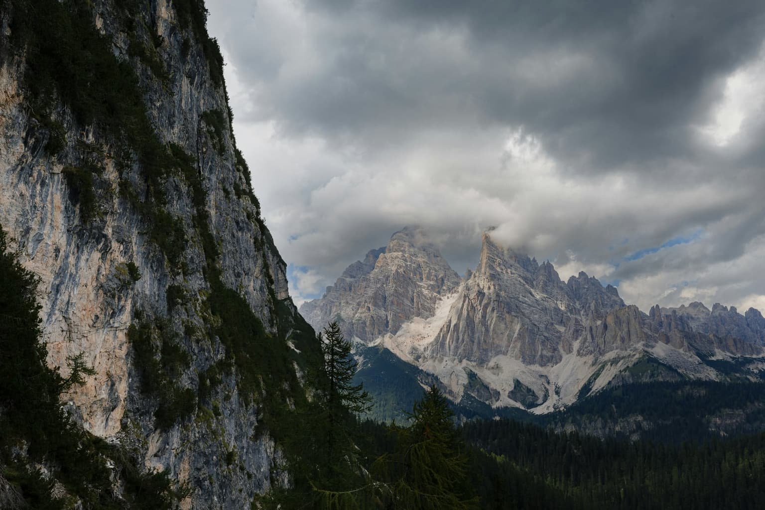Dark storm clouds pressing down on pale Dolomite spires, their summits already in cloud, above a shadowed cliff and dark forest