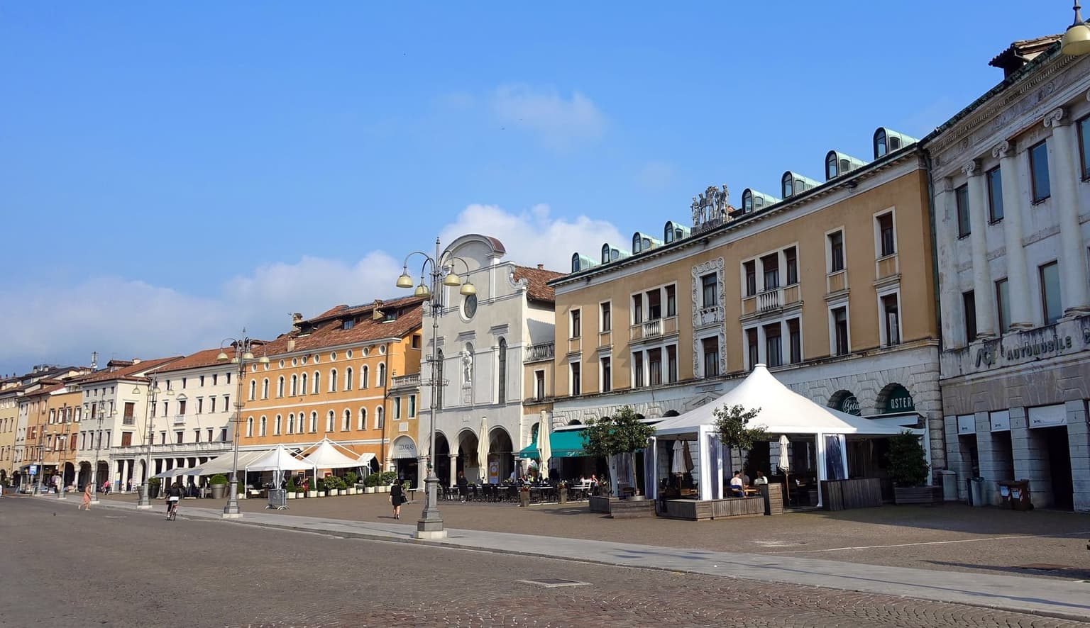Piazza dei Martiri in Belluno on a clear morning, a row of arcaded palazzi in ochre, white and cream with café umbrellas under the arches