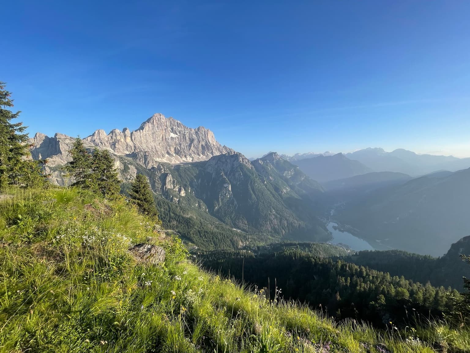 The pale limestone wall of the Civetta in early morning sun, with Lago di Alleghe and its village far below and a slope of wildflowers in front