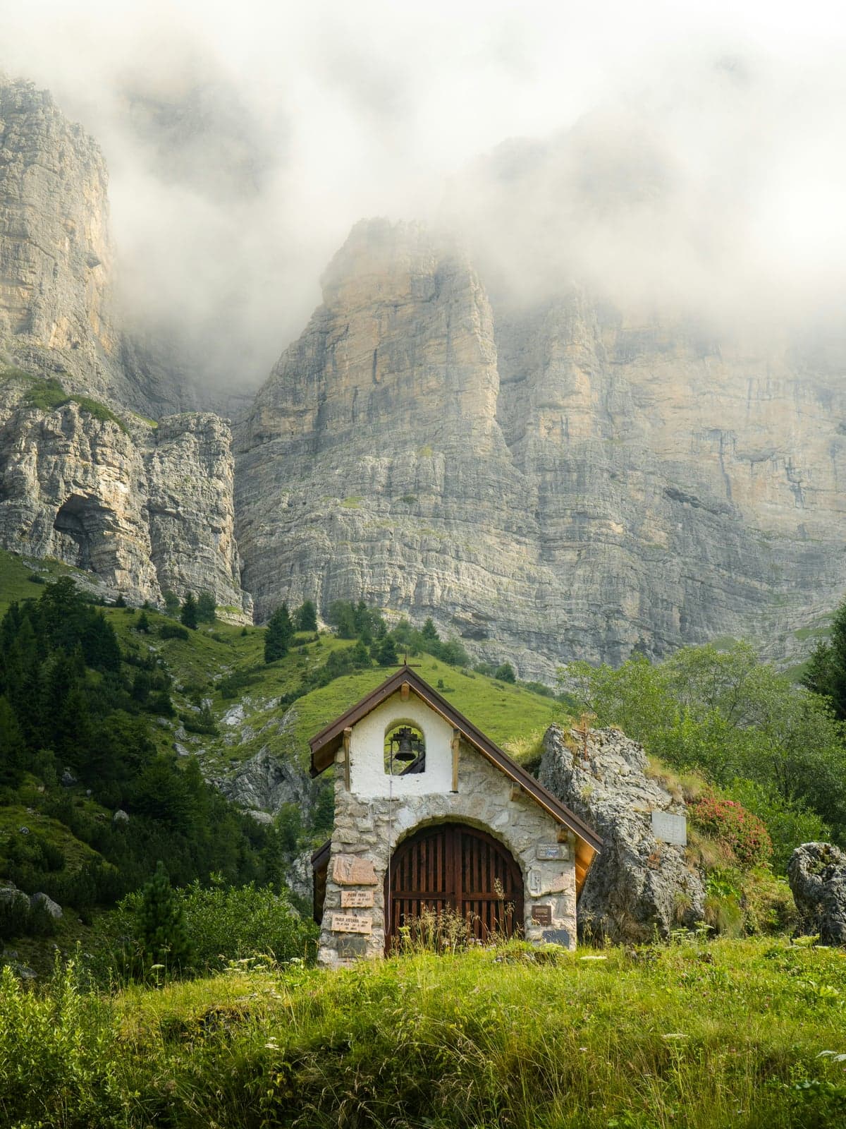 A small stone chapel with a bell stands on a green slope beneath sheer gray limestone walls whose tops disappear into cloud