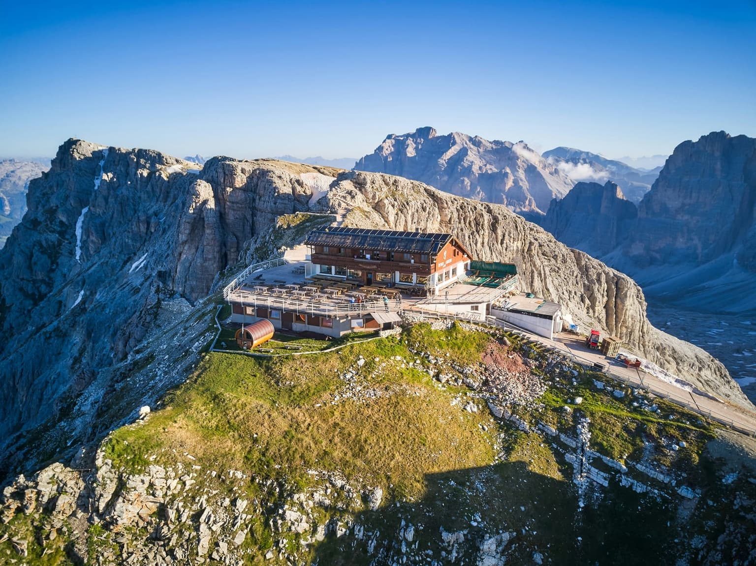 Aerial view of Rifugio Lagazuoi on its ridge in early sun, the wide terrace of tables in front of the hut and gray cliffs dropping away on both sides