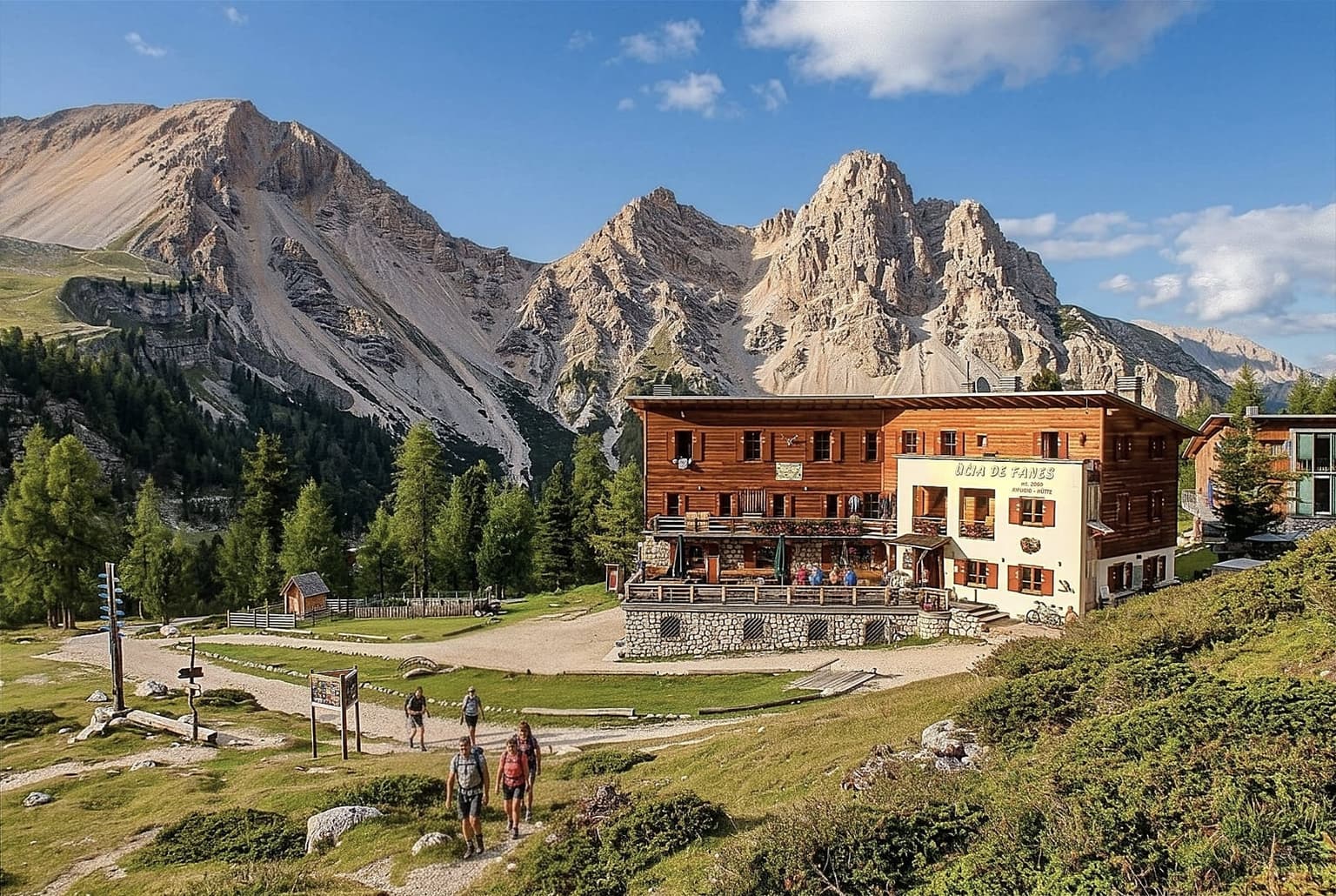Rifugio Fanes, a large timber hut with a white-painted wing signed Ücia de Fanes, 2,060 m, on a grassy pasture among larches, pale rocky peaks and long scree slopes behind it and a group of hikers walking up the gravel path in front