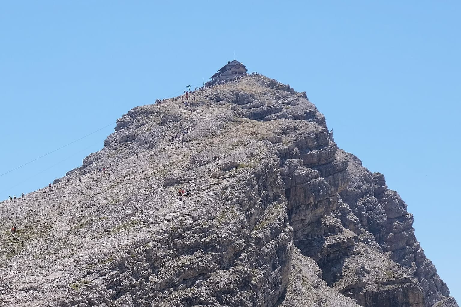 Rifugio Nuvolau perched on the very top of a bare limestone summit, hikers strung along the rock slabs climbing toward it and broken cliffs falling away on the right