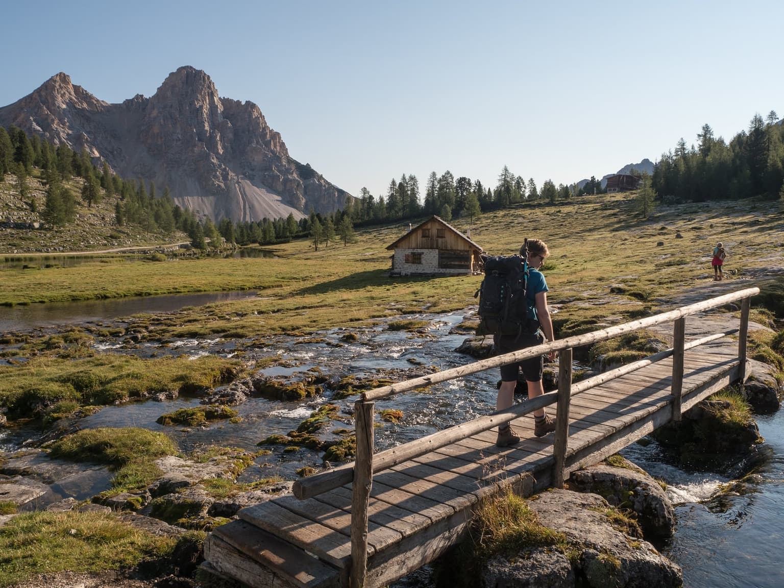 A hiker with a big trekking pack crosses a wooden footbridge over a clear stream on a high mountain pasture in early light, a small stone-and-timber hut and a pale rocky peak behind