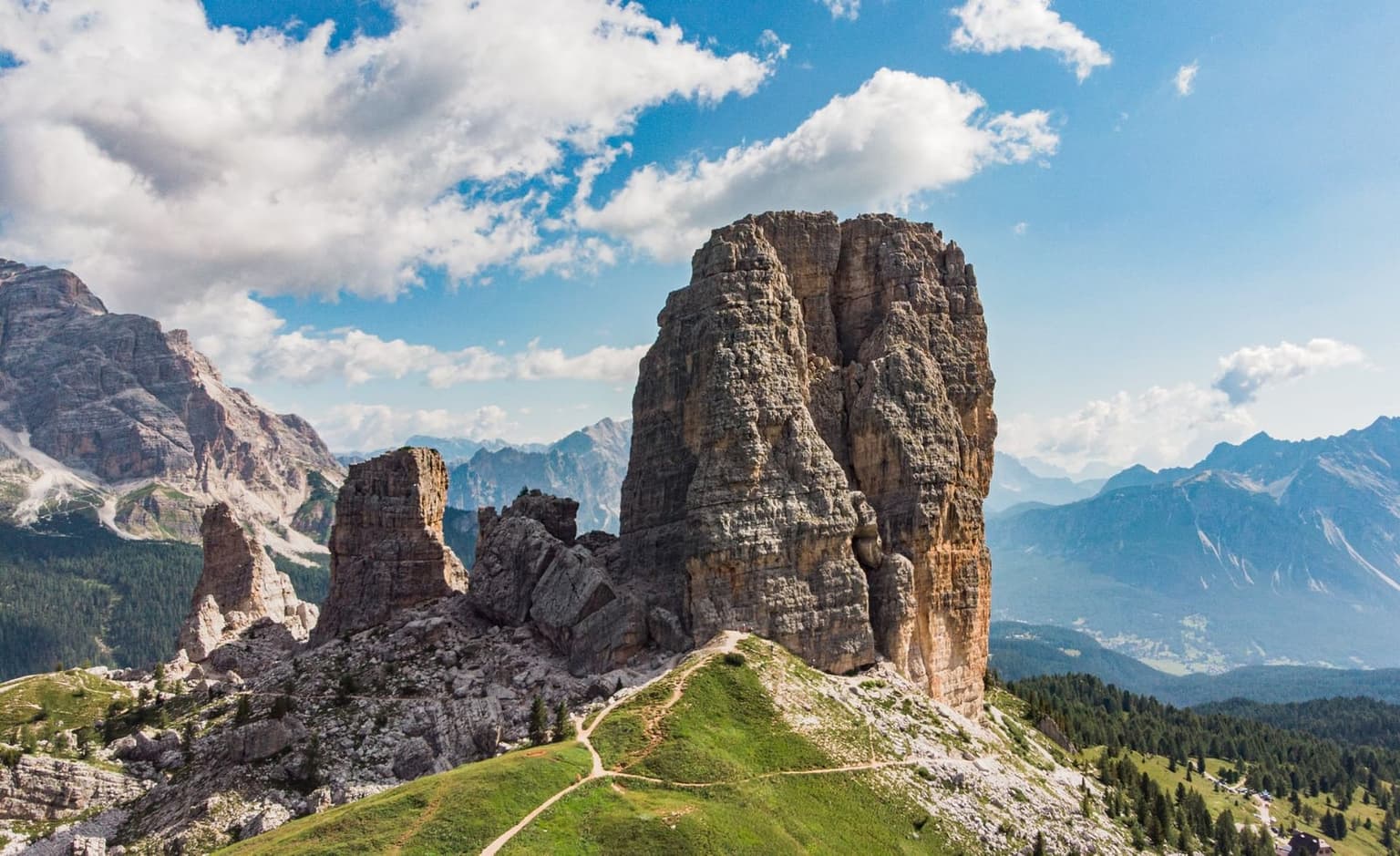 The five rock towers of Cinque Torri rising from a grassy ridge, hiking trails and small figures visible at their base, the Cortina valley beyond.
