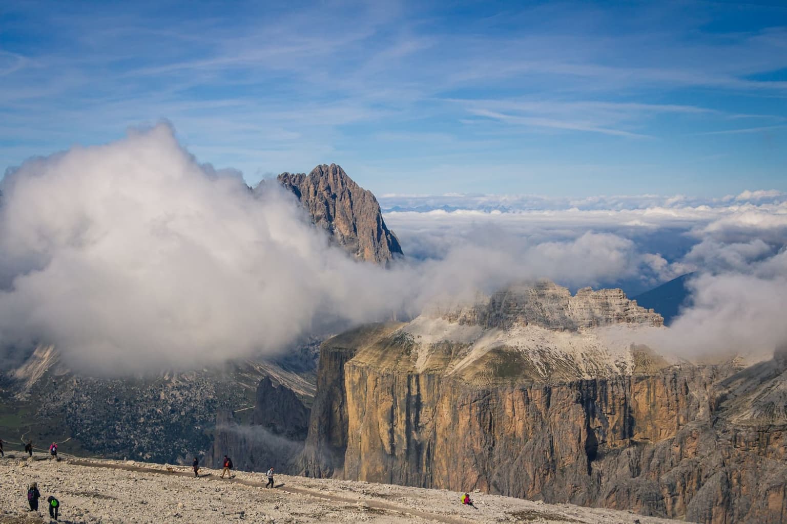 Hikers on a scree ridge above a sea of clouds, with layered Dolomite peaks and a flat-topped mountain visible in the distance.