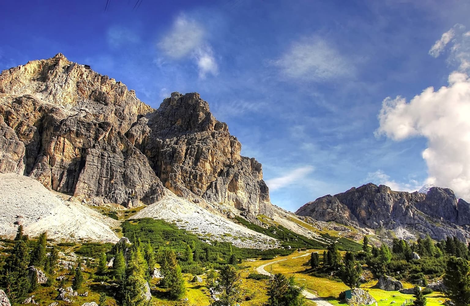 A rocky Dolomite ridge with a small structure visible near the summit and a trail winding through grass and scree below.