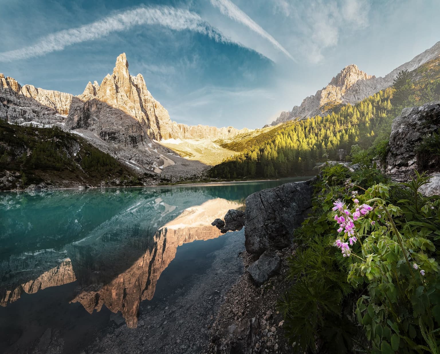 The turquoise water of Lago di Sorapis with a jagged Dolomite peak rising behind it and reflected in the surface, wildflowers in the foreground.