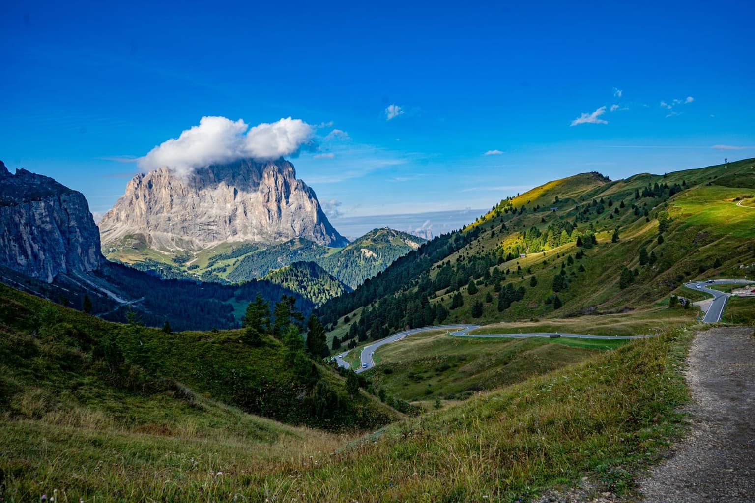 A paved mountain road winding through tight switchbacks with a few cars on it, a dramatic Dolomite peak rising behind.