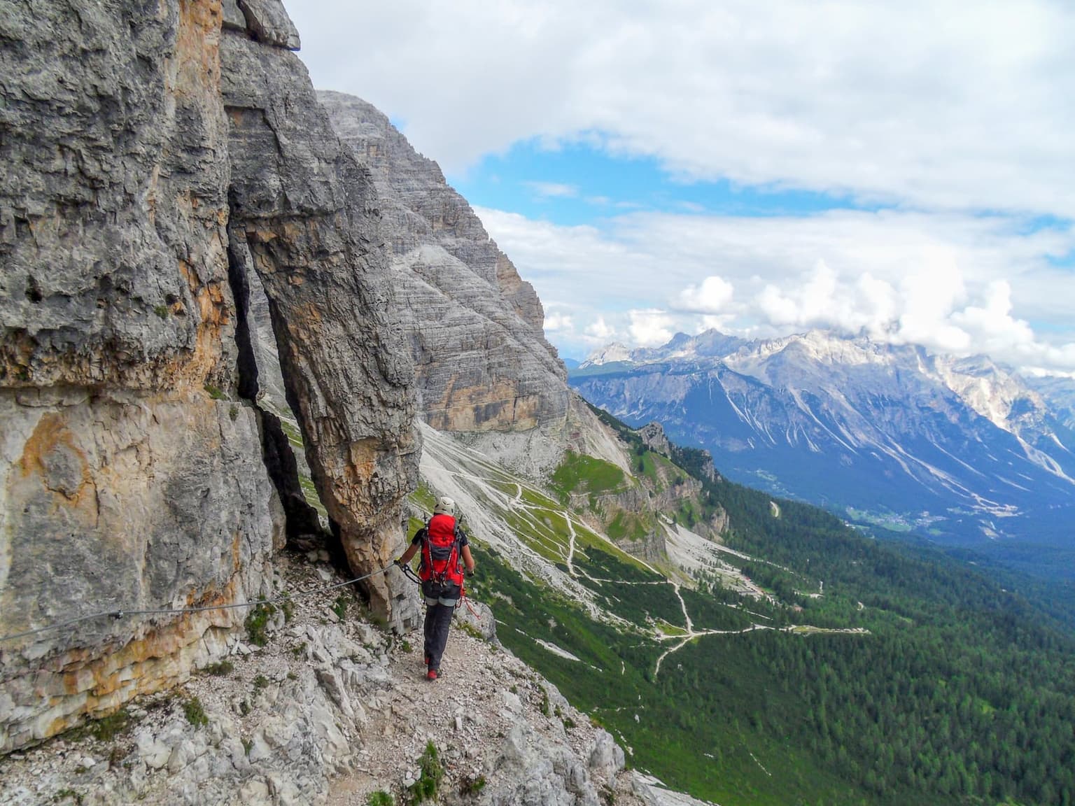 A hiker in a red backpack and white helmet clipped to a cable on a narrow via ferrata ledge, a steep drop and layered Dolomite peaks visible below.