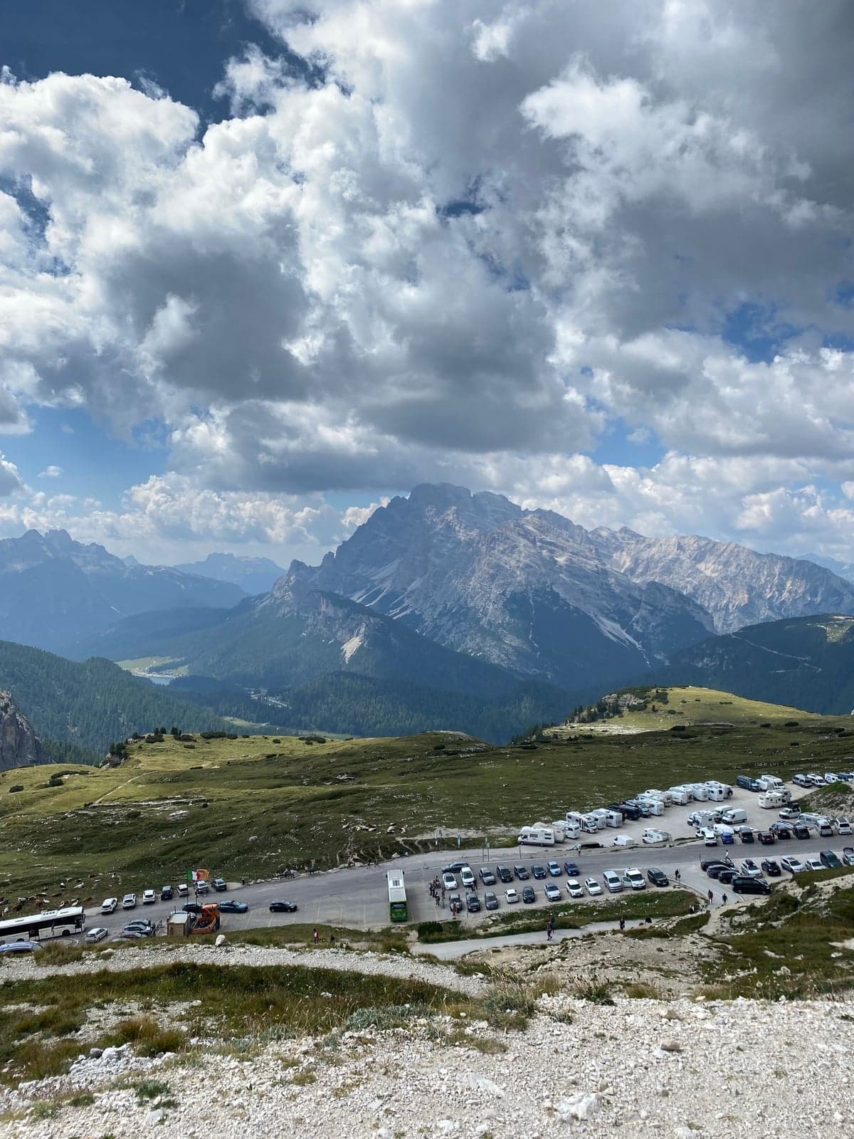 A mountain-pass parking lot packed with cars, campervans, and a tour bus under a partly cloudy sky, with snow-streaked Dolomite peaks and a small lake visible in the valley below