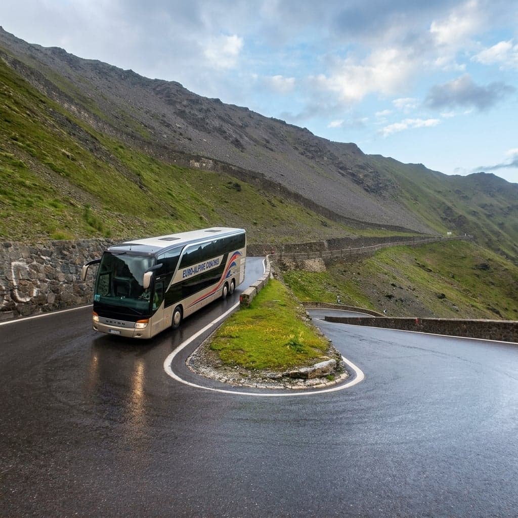 A large tour coach takes a tight hairpin turn on a narrow, wet mountain pass road, a stone wall on the inside of the curve and a steep green slope falling away on the outside