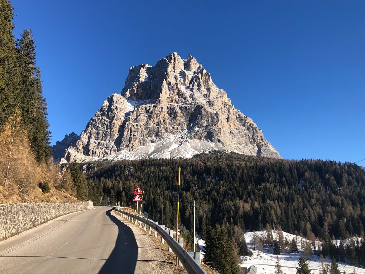 A two-lane mountain road curves along a stone retaining wall toward a towering, snow-dusted triangular Dolomite peak under a clear blue sky, guardrail and yellow hairpin-curve warning signs lining the roadside