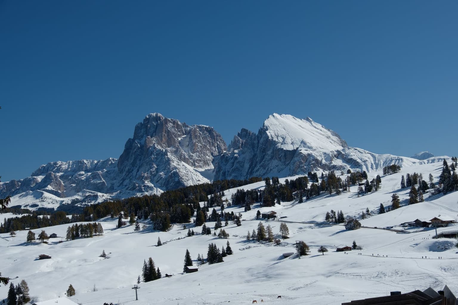 Walkers in single file on a groomed snow track across the Alpe di Siusi plateau, chairlift pylons and chalets nearby, with the Sassolungo and Sassopiatto peaks under a clear blue sky