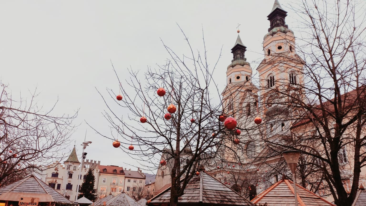 Wooden Christmas market stall roofs on Bressanone's cathedral square, red ornaments hanging from bare trees, the cathedral's twin towers behind on an overcast day