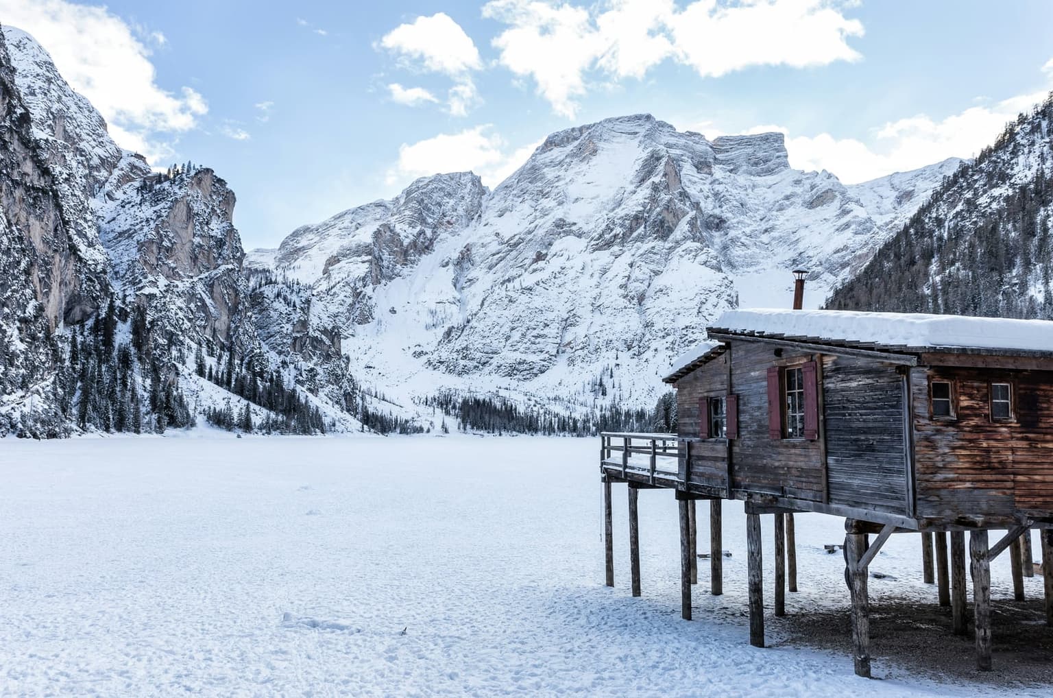 The frozen, snow-covered surface of Lago di Braies beside its wooden boathouse on stilts, the snowy Croda del Becco massif rising behind under scattered clouds