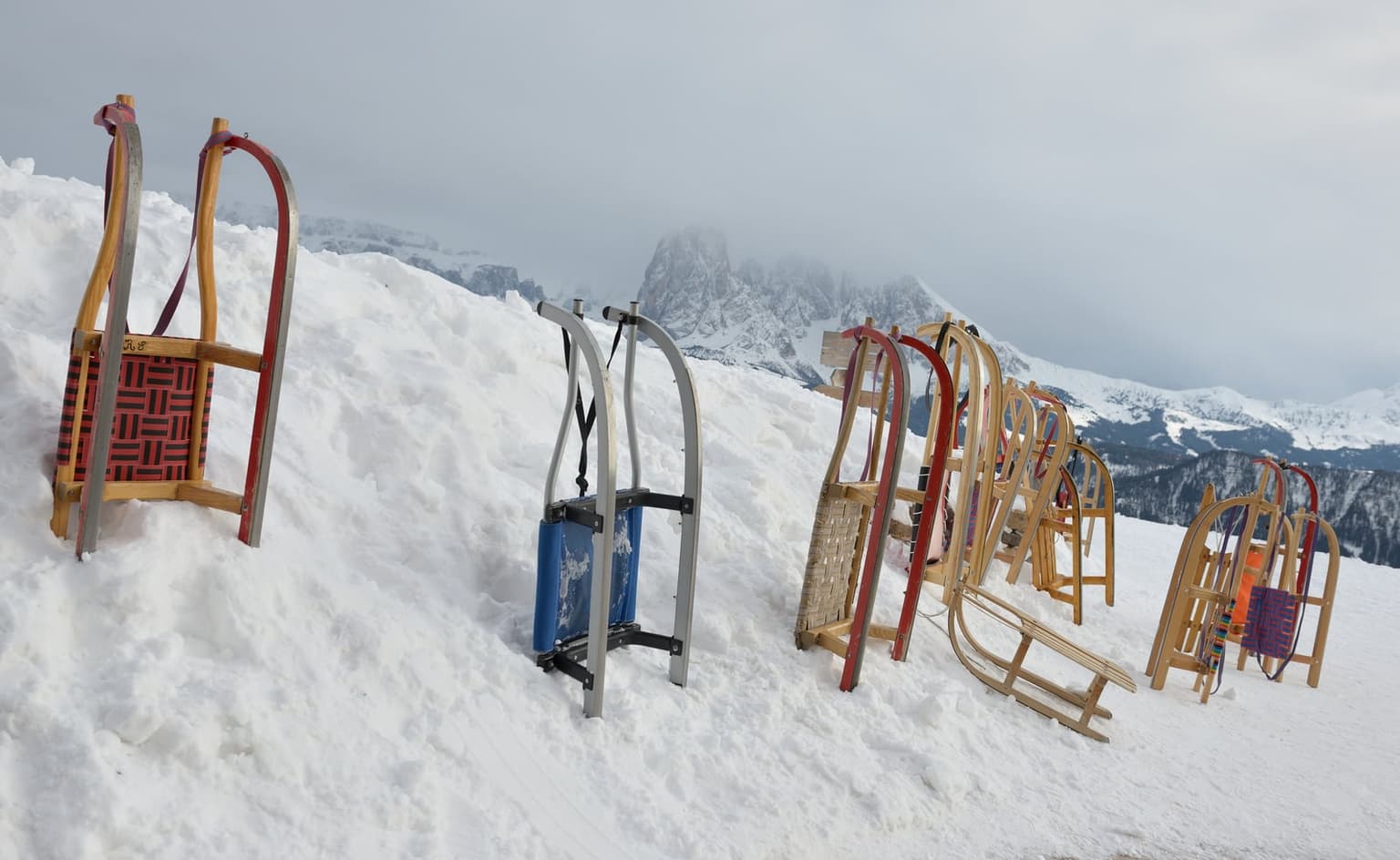 About a dozen wooden and metal sleds stuck upright in a snowbank at the top of a toboggan run, with the jagged Sassolungo group half-hidden in low cloud behind them